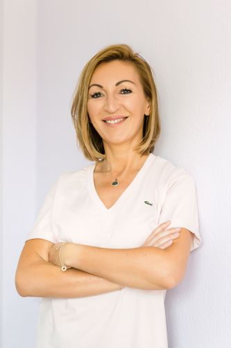 Porträt einer lächelnden Frau in heller Kleidung mit verschränkten Armen vor einer weißen Wand.